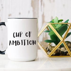 Könnte beinhalten: Weiße Keramiktasse mit schwarzem Henkel und Rand, mit dem Text "CUP OF AMBITION" in Schwarz. Daneben ein geometrisches goldenes Terrarium mit einer grünen Pflanze. Die Tasse steht auf einer weißen Oberfläche.