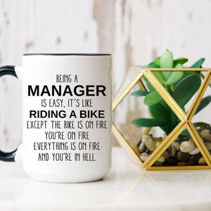 Puede incluir: Taza de cerámica blanca con asa y borde negros. La taza presenta el texto: "SER UN MANAGER es fácil, es como MONTAR EN BICICLETA, excepto que LA BICICLETA ESTÁ EN LLAMAS, TÚ ESTÁS EN LLAMAS, TODO ESTÁ EN LLAMAS Y ESTÁS EN EL INFIERNO."