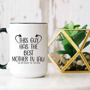 May include: White ceramic mug with a black handle and rim. The mug features the text "THIS GUY HAS THE BEST MOTHER IN LAW" in black script. A decorative geometric terrarium with a green plant sits beside the mug.