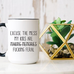 May include: White ceramic mug with a black handle and rim. The mug features the text "EXCUSE THE MESS MY KIDS ARE MAKING MEMORIES FUCKING FERAL" in black. A geometric terrarium with a faux plant is next to the mug.