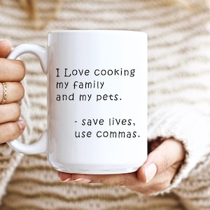 Puede incluir: Taza de cerámica blanca con el texto "I Love cooking my family and my pets. - save lives, use commas." La taza es sostenida por una persona que lleva un suéter beige y anillos dorados.