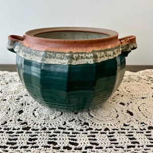 May include: A handcrafted ceramic bowl with a dark green exterior and a brown rim. The bowl has two handles and a white and green band around the middle. The bowl is sitting on a white crocheted doily.