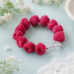Op de afbeelding: Een armband met een reeks realistische, felrode, framboosvormige kralen in een cirkelvormig patroon. De armband wordt vastgemaakt met een gepolijste, zilverkleurige knevelsluiting, waaraan een kleine, gedetailleerde metalen vogelbedel is bevestigd.