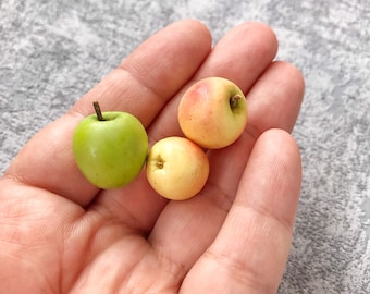 May include: Three miniature apples, one green and two yellow-orange, are held in a person's hand. The apples are small and round, resembling miniature fruit.