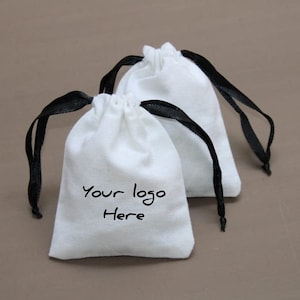 Puede incluir: Dos bolsas de algodón blanco con cordón de ajuste y cintas negras. La bolsa en primer plano tiene el texto "Your logo Here" impreso en la parte delantera.