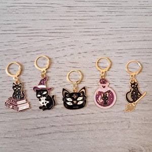 May include: Five gold charms with black cat designs. Each charm features a different cat-themed design, including a cat on books, a cat with a witch hat, a cat with multiple eyes, a cat in a potion bottle, and a cat on a broom.