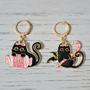 Op de afbeelding: Een paar goudkleurige oorringen met zwarte kattenontwerpen. Eén kat houdt een roze en wit snoepje vast, de andere heeft een vleermuisvleugel en een roze en witte zuurstok. De oorbellen hebben zwarte emaille en roze accenten.