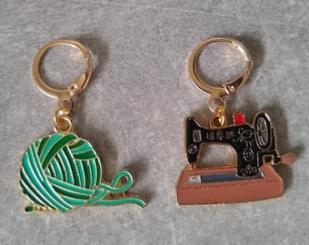 May include: A pair of gold-colored hoop earrings. One earring features a green and teal yarn ball charm, and the other has a black and brown sewing machine charm. The charms are detailed with enamel.