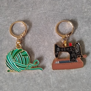 Op de afbeelding: Een paar goudkleurige oorringen. Eén oorring heeft een groene en blauwgroene bol garen bedel, de andere heeft een zwart en bruin naaimachine bedel. De bedels zijn gedetailleerd met emaille.