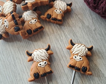 May include: A collection of brown and white Highland cow-shaped embellishments, with black horns and details, scattered on a light gray surface. A pair of red-handled scissors and a ball of burgundy yarn are also present. Some of the cows are attached to silver pins.