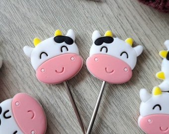 May include: Close-up of several cow-shaped craft tools with stainless steel stems. The cows have white faces, black eyes, and pink noses and ears. Yellow horns and a smiling mouth complete the design. A pair of scissors and a ball of burgundy yarn are in the background.