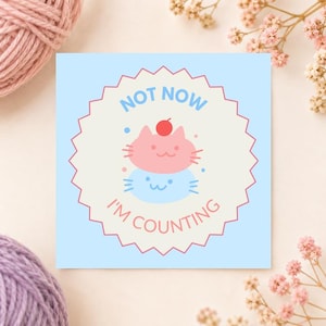 Può includere: Un adesivo quadrato con sfondo azzurro e un cerchio color crema dai bordi smerlati. All'interno ci sono due facce di gatto sovrapposte, una rosa e una blu, con una ciliegia rossa in cima. Il testo recita NOT NOW I'M COUNTING in blu e rosa.