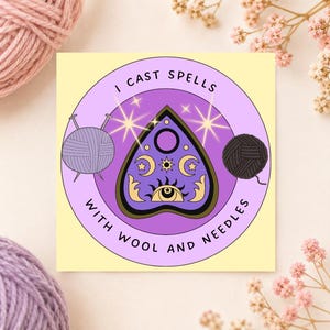 Könnte beinhalten: Ein runder Aufkleber mit hellviolettem Hintergrund und einer zentralen violetten Planchette, die mit Sonne, Mond und Auge verziert ist. Der Text I CAST SPELLS WITH WOOL AND NEEDLES umgibt die Planchette, flankiert von zwei Wollknäueln.