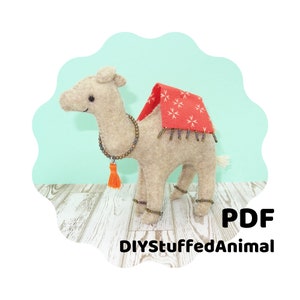 May include: A beige felt camel plush toy with a red and white patterned saddle and beaded trim. The camel is wearing a beaded necklace with a tassel. The toy is on a light wood surface. DIYStuffedAnimal PDF.