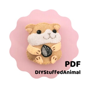 Könnte beinhalten: Ein braunes und weißes Filzhamster, der eine graue Filznuss hält. Der Hamster sitzt auf einem rosa Hintergrund. Der Text "DIYStuffedAnimal" und "PDF" befindet sich unter dem Hamster.