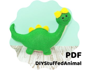 May include: A green felt dinosaur with yellow accents and a yellow bow. The dinosaur has a smiling face and is standing on a white wooden surface. PDF DIYStuffedAnimal