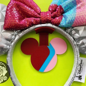 Op de afbeelding: Zilveren en grijze Mickey Mouse orenband met een roze, blauwe en witte pailletten strik. De hoofdband heeft een rode, roze en blauwe Mickey Mouse silhouet bedel. De hoofdband heeft een Disney Parks logo label.