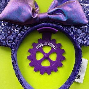 May include: Purple sequined Minnie Mouse ears with a purple bow and a purple plastic gear with the text "Carousel of Progress" on it.