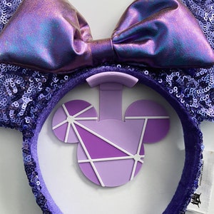 May include: Purple sequined ear headband with a large iridescent bow. The headband features a geometric Mickey Mouse design in shades of purple and white. A fun accessory for theme park enthusiasts.