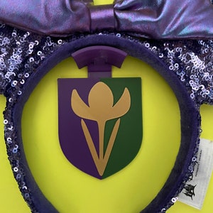 May include: Purple and iridescent sequined Minnie Mouse ears with a purple and green flower charm attached. The charm has a gold outline. The ears have a white tag attached with the text "AUTHENTIC DISNEY PARKS MERCHANDISE".