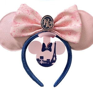 May include: Pink Minnie Mouse ears with a blue headband. The bow is pink with a white and pink floral pattern. The ears have a pink silhouette of the Eiffel Tower and a building with a Parisian skyline. The bow has a gold charm with the letter "R" on it.