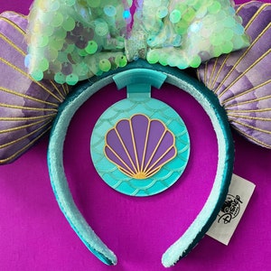 May include: Purple and teal Minnie Mouse ears with a green sequin bow and a purple seashell charm. The ears are shaped like seashells and have gold trim. The charm is attached to a teal headband with a Disney logo.