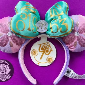 May include: A pair of purple and pink Minnie Mouse ears with a turquoise bow featuring the text "50" and "63" in gold. The bow has a small, round, silver charm with a design of a castle and a clock. The ears are attached to a purple headband with a white tag featuring a gold design of a castle and a clock.