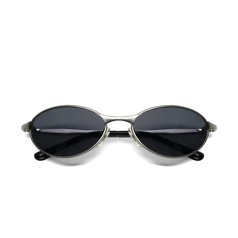 May include: Oval-shaped sunglasses with dark lenses and a silver metal frame. The sunglasses have a sleek, minimalist design, with a thin bridge and temples. The lenses appear to be tinted, providing shade and protection from the sun.