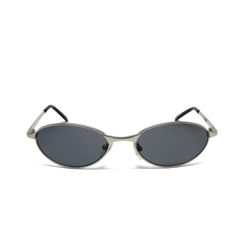 May include: Oval-shaped sunglasses with a silver metal frame and dark gray lenses. The sunglasses have a sleek, minimalist design with thin temples and a classic, retro aesthetic. The lenses are slightly curved.