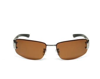 May include: Sunglasses featuring brown tinted lenses and a silver-toned metal frame. The design includes a thin bridge, adjustable nose pads, and dark-colored temples that curve at the ends. The lenses are semi-rimless, held by silver metal accents.