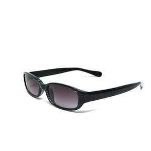 Puede incluir: Gafas de sol de plástico negro con lentes gris oscuro. Las gafas tienen una forma rectangular y un marco delgado.