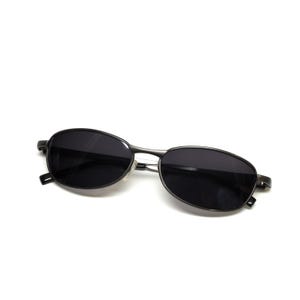 May include: Oval-shaped sunglasses with dark lenses and a thin, metallic frame. The sunglasses are a dark gray color and have a minimalist design, suitable for various styles. The lenses appear to be tinted, offering protection from sunlight.