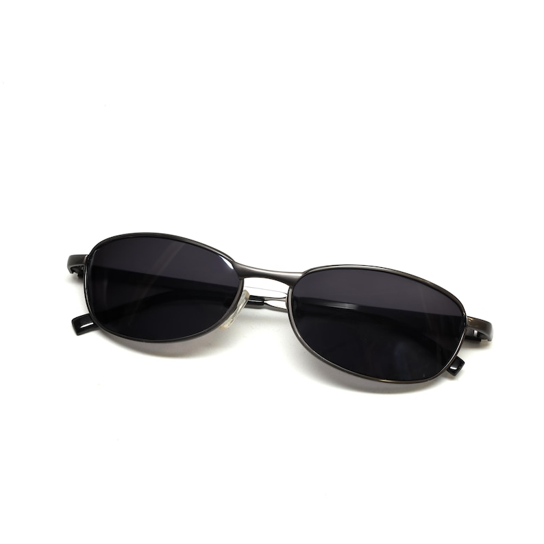 May include: Oval-shaped sunglasses with dark lenses and a thin, metallic frame. The sunglasses are a dark gray color and have a minimalist design, suitable for various styles. The lenses appear to be tinted, offering protection from sunlight.
