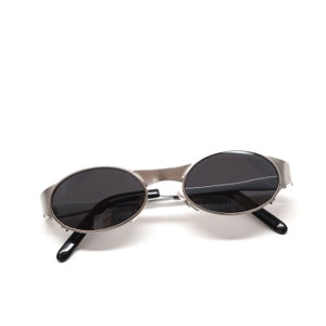 Puede incluir: Gafas de sol ovaladas plateadas con lentes negros. Las gafas est&aacute;n sobre una superficie blanca.