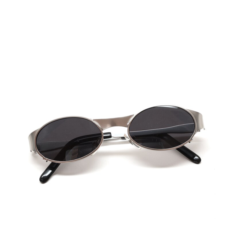 Puede incluir: Gafas de sol ovaladas plateadas con lentes negros. Las gafas est&aacute;n sobre una superficie blanca.