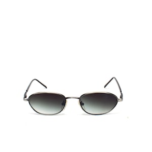 Pode incluir: Óculos de sol ovais com lentes degradê. As armações são de cor metálica escura e as hastes são finas e elegantes. As lentes transitam de um tom escuro na parte superior para um tom mais claro na parte inferior.
