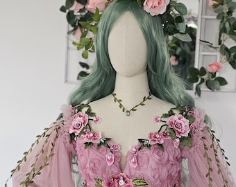 May include: A pink tulle gown with floral embellishments and long, sheer sleeves. The bodice features intricate floral detailing and a plunging neckline. The dress is adorned with pink flowers and green vines, creating a whimsical, fairy-tale aesthetic.