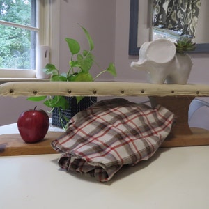 Könnte beinhalten: Ein vintage Holzbügelbrett mit gepolsterter Oberseite und einer weißen Keramik-Elefantenfigur mit einer Sukkulentenpflanze darauf. Ein roter Apfel und ein karierter Stoff liegen auf dem Tisch neben dem Bügelbrett.