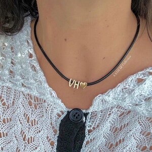 May include: A gold-coloured necklace featuring the initials "VH" and a heart charm. The letters and heart are attached to a thin, black cord. The necklace is worn against a white and black patterned top.