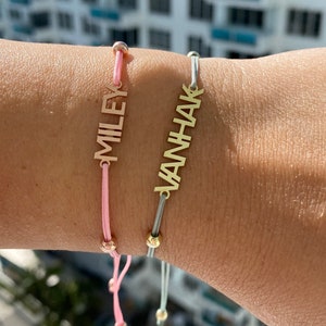 Peut inclure: Deux bracelets personnalisés avec les noms "MILEY" et "VANHAK" gravés dessus. Les bracelets sont faits de breloques en métal couleur or et de cordons réglables. Le bracelet de gauche a un cordon rose et le bracelet de droite a un cordon gris.