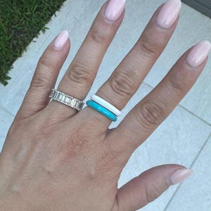 May include: A hand displaying several rings. One ring features a row of rectangular-cut stones. Two other rings are stacked, one white and one turquoise. The nails are painted with a light pink polish.