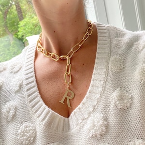 Op de afbeelding: Een gouden ketting met een grote gouden letter 'R' hanger. De ketting wordt gedragen door iemand die een witte trui draagt.