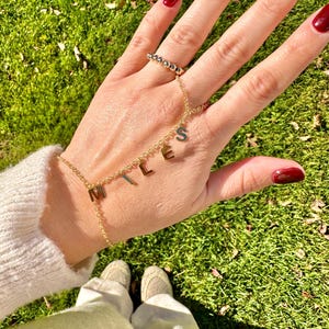 Könnte beinhalten: Ein goldenes Kettenarmband mit dem Wort "MILES" in Charms. Das Armband wird am Handgelenk getragen, mit einem goldenen Perlenring an derselben Hand.