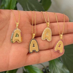 May include: Four gold necklaces with letter 'A' pendants. Each pendant is outlined with colorful gemstones. The necklaces are on a hand.