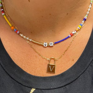 May include: Two gold necklaces with a padlock charm on the bottom. The top necklace is made of colorful beads and the bottom necklace is a simple chain with a padlock charm. The padlock charm has the letter "V" on it.