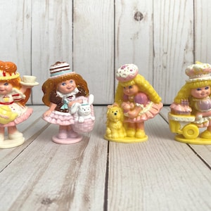 May include: Four vintage miniature figurines of girls with blonde and red hair, wearing dresses and hats. One holds a tea cup, another a basket with a bunny, and the others have a dog and a cake cart. The figurines are on a light wood surface.