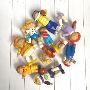 Pode incluir: Uma coleção de bonecas Cabbage Patch Kids vintage, cada uma vestida com roupas coloridas. Algumas bonecas estão usando uniformes de futebol americano, e outras roupas casuais. As bonecas estão em uma superfície de madeira branca.