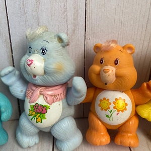 Puede incluir: Dos figuras de oso de pl&aacute;stico. El oso de la izquierda es azul claro con una bufanda rosa y una mancha blanca en el vientre con una rosa rosa. El oso de la derecha es naranja con una mancha blanca en el vientre con dos flores amarillas sonrientes.