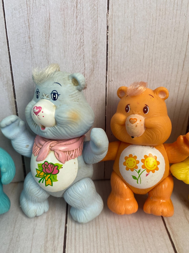 Puede incluir: Dos figuras de oso de pl&aacute;stico. El oso de la izquierda es azul claro con una bufanda rosa y una mancha blanca en el vientre con una rosa rosa. El oso de la derecha es naranja con una mancha blanca en el vientre con dos flores amarillas sonrientes.