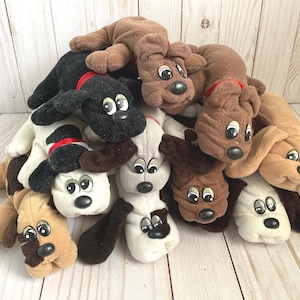 Puede incluir: Un montón de juguetes de peluche con forma de perro en varios colores, incluyendo marrón, negro y blanco. Cada perro tiene ojos grandes y expresivos y orejas caídas. Algunos tienen collares rojos.